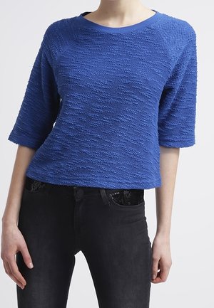 Blauwe gebreide trui met driekwartmouwen en een ronde hals, met een licht cropped ontwerp. Draagt bij zwarte spijkerbroek.