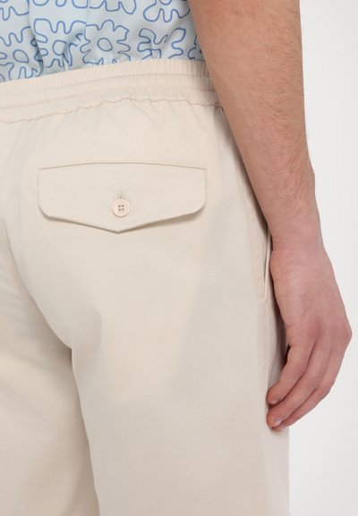 Pantaloni bej din bumbac cu talie elastică, având o pamblică la spate cu închidere prin buton. Textură netedă, design simplu.