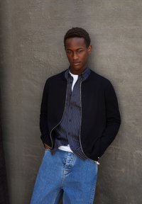 Zwarte zip-up bomberjack over een gestreepte overhemden met knopen, gecombineerd met lichtblauwe denimjeans. Gladde textuur, relaxed fit.