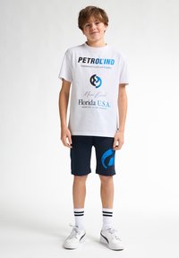 Bílý tričko s modro-černým potiskem, doplněné námořnickými kraťasy s velkým modrým logem. Bílá sneakers a pruhované ponožky dokončují outfit.