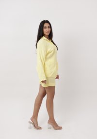 Mujer de perfil vistiendo una camisa amarilla holgada y pantalones cortos a juego con sandalias transparentes de tacón alto sobre un fondo blanco.