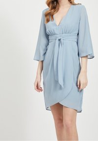 Žena v světle modrých šatech s přehozem, tříčtvrtečním rukávem a páskem kolem pasu, stojící proti jednoduchému světlému pozadí.