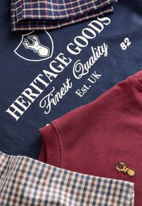 Warstwowo ułożone ubrania obejmują granatową koszulę z białym tekstem "HERITAGE GOODS Finest Quality Est. UK 82", bordowy element garderoby i dwa tkaniny w kratę.