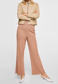 Žena oblečená v béžovém zkráceném koženém saku, odpovídajících pletených širokých kalhotách a bílých špičatých slingback podpatcích, stojí se zkříženýma rukama.