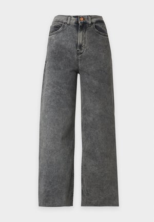 Mujer con chaqueta crema con cremallera, top negro, jeans gris de pierna ancha y botas negras de tacón, de pie con las manos en los bolsillos.