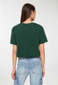 Donna con lunghi capelli castani che indossa una maglietta verde scuro a maniche corte e jeans a vita alta blu chiaro con tasche a toppa, girata di spalle.