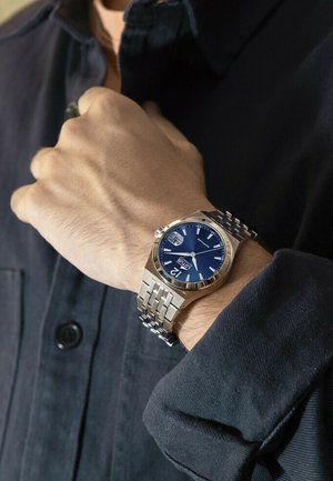 Silberner Edelstahl-Armbanduhr mit blauem Zifferblatt, Datumsanzeige und silbernem Gliederarmband, getragen an einer Hand vor einem dunklen Hemd-Hintergrund.