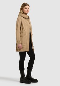 Femme debout portant un manteau à capuche beige, des leggings noirs et des bottes noires épaisses, sur un fond clair uni.