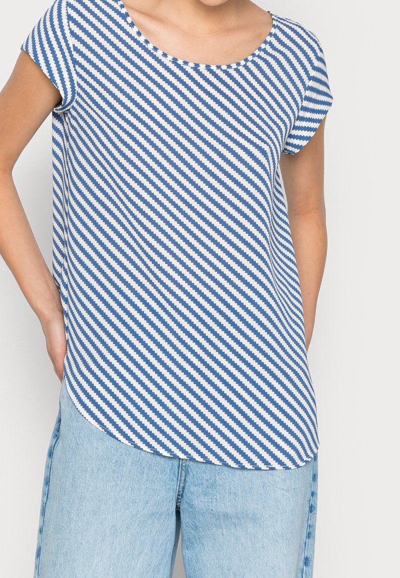 Personne portant un haut à manches courtes rayé bleu et blanc en diagonale, associé à un jean en denim bleu clair, main posée sur la hanche.