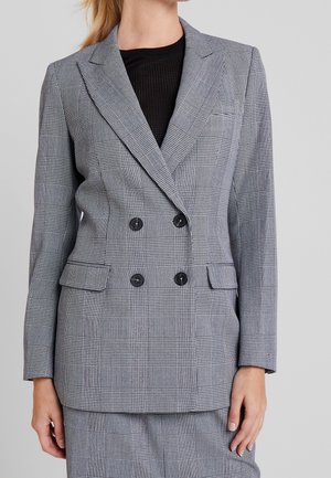 Kvinde iført grå ternet dobbeltknappet blazer med sorte knapper og matchende nederdel, stående mod en ensfarvet baggrund.