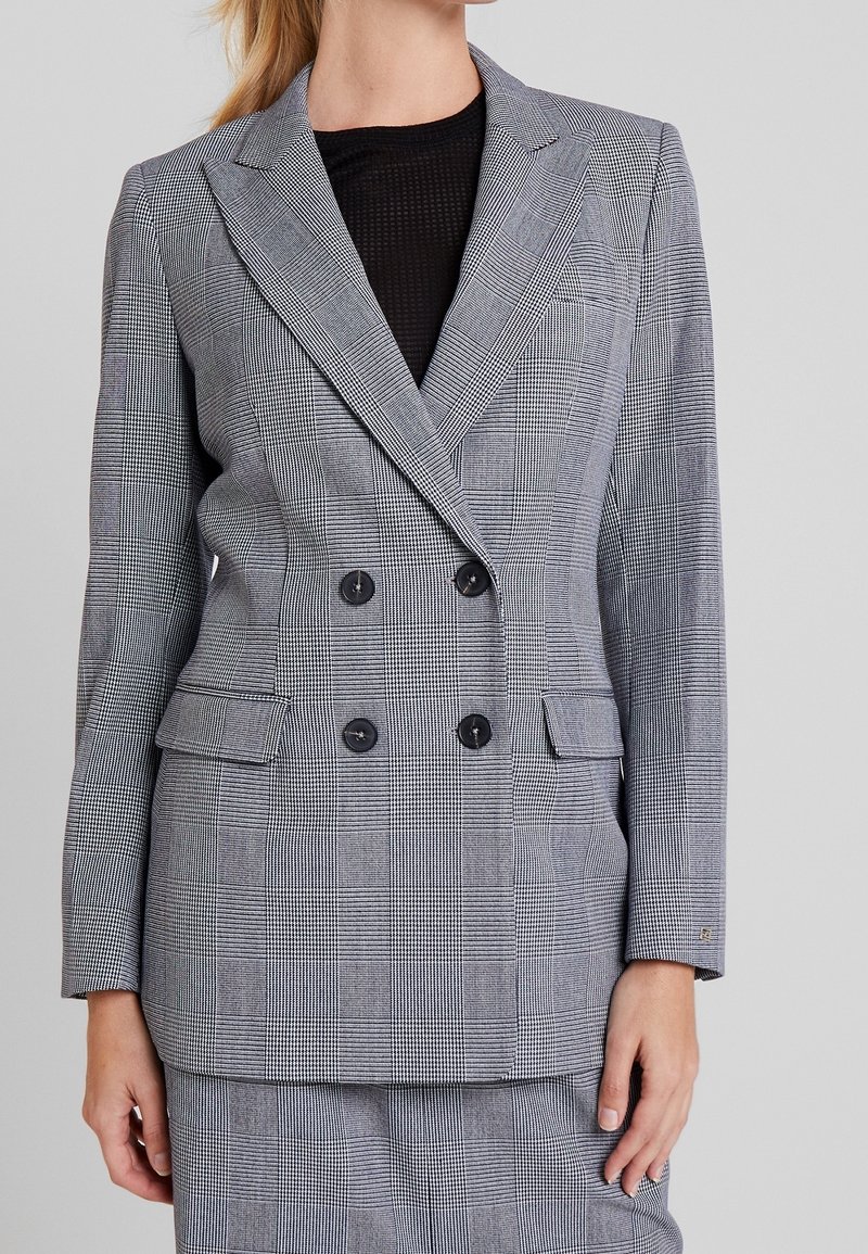 Femme portant un blazer croisé à carreaux gris avec des boutons noirs et une jupe assortie, debout devant un fond uni.