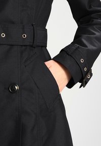 Black trench coat featuring a smooth fabric, a belted waist, a button closure, and gold-tone hardware accents on cuffs and pockets.