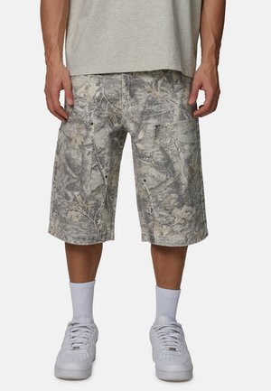 Mężczyzna w szarych szortach moro typu cargo, białych skarpetach do kostek i białych trampkach, stojący z luźno opuszczonymi rękami.