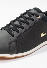 Svart låg sneaker med svarta snören, texturförsedda sidor, vit sula och liten guldig krokodil-logotyp på sidan.