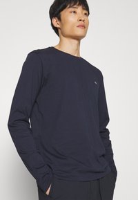 T-shirt à manches longues navy en coton doux ; col rond, coupe décontractée, avec un logo subtil sur la poitrine et des manches fuselées.