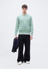 Jeune homme portant un pull vert menthe, des baskets blanches et un jean noir large, tenant une veste bleu marine pliée devant un fond blanc uni.