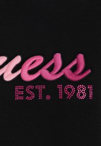 Testo ricamato in rosa e con strass che recita "Guess EST. 1981" su uno sfondo di tessuto nero.