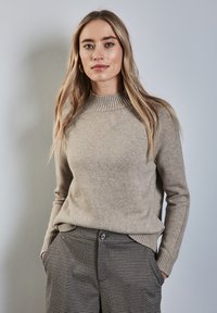 Beige gebreidde trui met ribbels aan de halslijn, gecombineerd met geruite broek met zijzakken en een knoopsluiting. Zachte textuur.