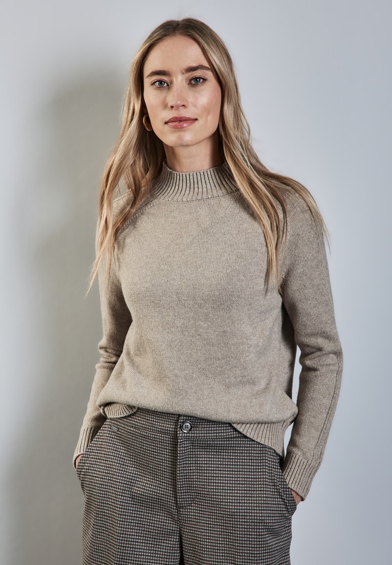 Beige gebreidde trui met ribbels aan de halslijn, gecombineerd met geruite broek met zijzakken en een knoopsluiting. Zachte textuur.