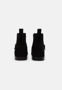 Ett par svarta mocka ankelstövlar med platta klackar, bakre dragremmar och sidobucklar, visade bakifrån mot enbart vit bakgrund.