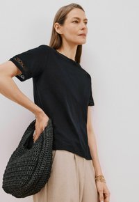 T-shirt nero in cotone con rifiniture in pizzo alle maniche, abbinato a una borsa nera tessuta e strutturata e pantaloni beige chiaro.