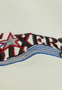 Bestickter cremefarbener Stoff mit dem Wort "BOXERS" in Schwarz, roten Akzenten und einem blauen Wellenmuster darunter. Texturierte Oberflächenstruktur sichtbar.