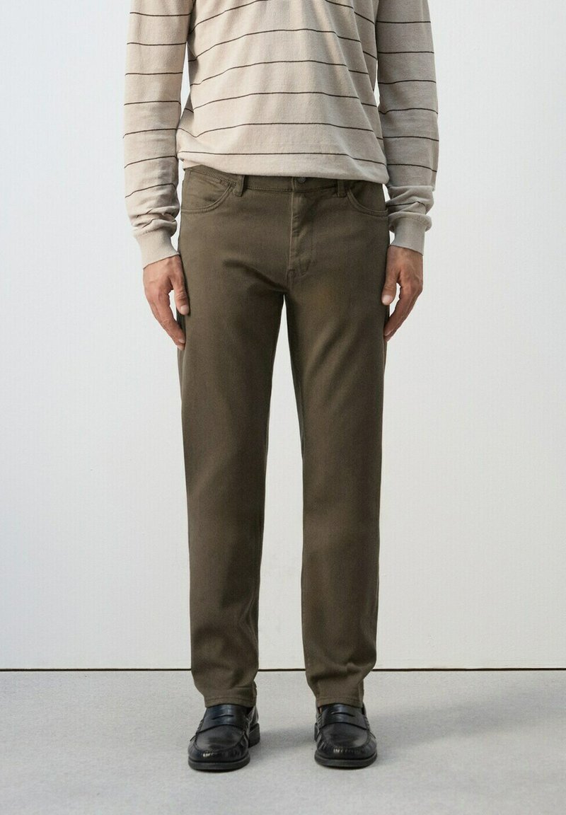 Mann trägt olivgrüne, gerade geschnittene Hose, beige langärmliges gestreiftes Hemd und schwarze Lederloafer, steht vor einem neutralen Hintergrund.