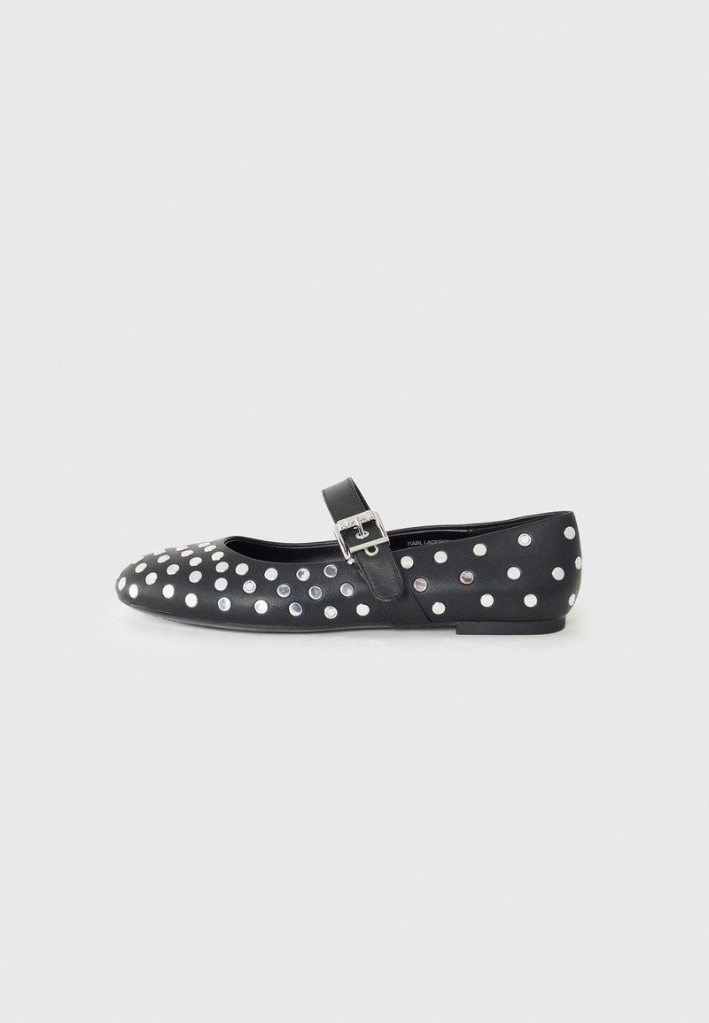 Chaussure plate Mary Jane noire avec des clous ronds argentés et une bride à boucle argentée sur un fond gris clair.