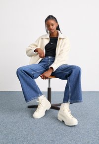 Giacca bianca foderata in pile, top nero aderente, jeans blu a vita alta e stivali larghi crema. Modella seduta su uno sgabello di legno su uno sfondo semplice.