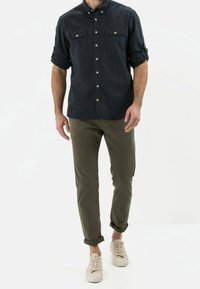 Dunkelblaues Button-Up-Hemd mit zwei Brusttaschen, hochgekrempelte Ärmel. Olivgrüne Chinos, am Knöchel umgeschlagen. Helle beigefarbene Sneaker.