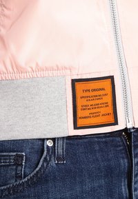 Giacca da volo bomber in rosa chiaro, con orlo a coste grigio e un'etichetta arancione con testo nero. Zip metallica e jeans di denim visibili.