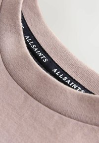 Beežist puuvillast T-särgi krae, millel on sile tekstuur. Kaela sees on nähtav must silt valge tekstiga "ALLSAINTS".