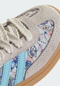 Gros plan sur une basket en daim beige, avec un tissu à motif floral, des bandes bleu clair, des lacets épais couleur crème et une semelle en caoutchouc naturel.