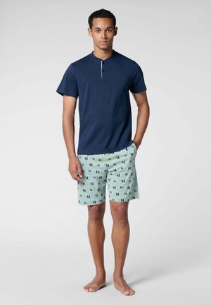 Uomo in piedi a piedi nudi che indossa una maglia henley manica corta blu navy e pantaloncini verde chiaro con motivi di pesci scuri.