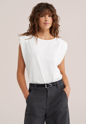 Femme aux cheveux bouclés porte un haut blanc sans manches avec des épaules rembourrées, rentrée dans un pantalon gris foncé avec une ceinture noire, mains dans les poches.