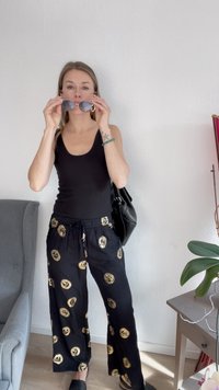 Svart tank top, svarte bukser med gull sirkulære mønstre, holder solbriller, står innendørs nær en plante og møbler.