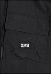Coupe-vent noir avec une fermeture éclair avant, un tissu texturé et une grande poche à rabat avec le logo Urban Classics en blanc.