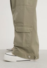 Pantalons cargo kaki avec poche latérale, détails cousus, ourlet retroussé et bouton-pression argenté. Portés avec des baskets blanches à bord noir.