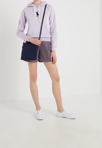 Felpa viola chiaro con tasca frontale e logo, abbinata a pantaloni corti grigio scuro. Regge una borsa a tracolla blu navy; indossa sneakers bianche.