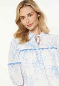 Blusa bianca con motivo floreale blu, dotata di colletto, dettaglio a fiocco sul collo e accentuazioni in pizzo blu su maniche e corpetto. Maniche lunghe e svasate.