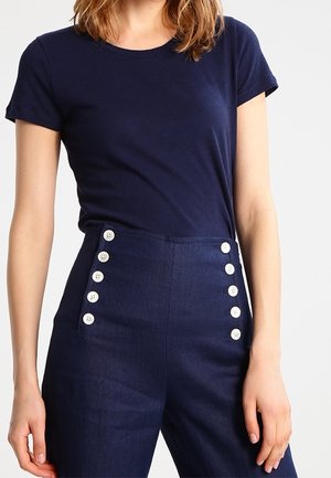 Kobieta ubrana w granatową koszulkę z krótkim rękawem oraz granatowe spodnie z wysokim stanem z dwoma pionowymi rzędami białych guzików z przodu.