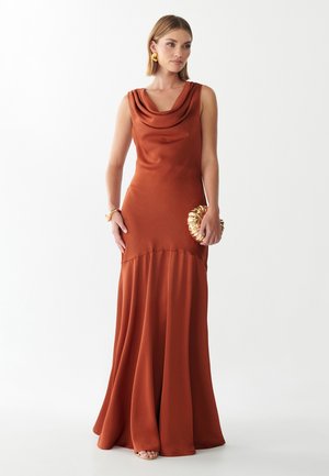 Vrouw in een roestkleurige, mouwloze satijnen jurk met een gouden, getextureerde clutch, afgewerkt met gouden oorbellen en armband, staand op een witte achtergrond.