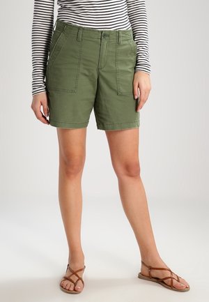 Person trägt grüne knielange Shorts, gestreiftes Langarmshirt und braune flache Riemchensandalen und steht vor einem schlichten hellen Hintergrund.