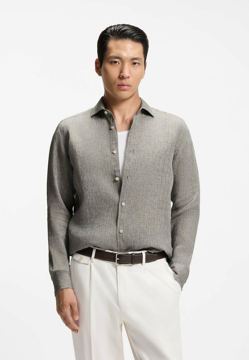 Homme portant une chemise gris clair à boutons sur un sous-vêtement blanc rentré dans un pantalon blanc avec une ceinture marron foncé, les mains dans les poches.
