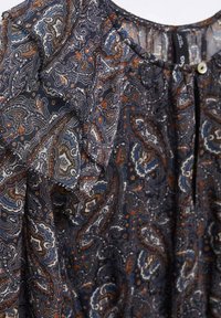 Blouse bleu foncé avec un motif paisley complexe en blanc, marron et bleu, avec une manche à volants et un petit bouton blanc près de l'encolure.