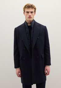 Cappotto doppiopetto in lana blu navy con interno strutturato, rever a incavo e quattro bottoni frontali, abbinato a un maglione nero.