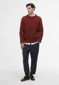 Άνδρας που φοράει κόκκινο και καφέ ριγέ πλεκτό πουλόβερ, λευκό πουκάμισο από κάτω, ναυτικό μπλε παντελόνι και μαύρα δετό παπούτσια, στέκεται και κοιτάζει δεξιά.