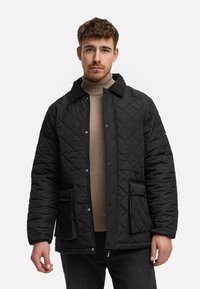 Zwarte gewatteerde jas met een kraag, drukknoopsluiting, twee voorkruizen en een zachte textuur, gedragen over een tan coltrui en donkere jeans.