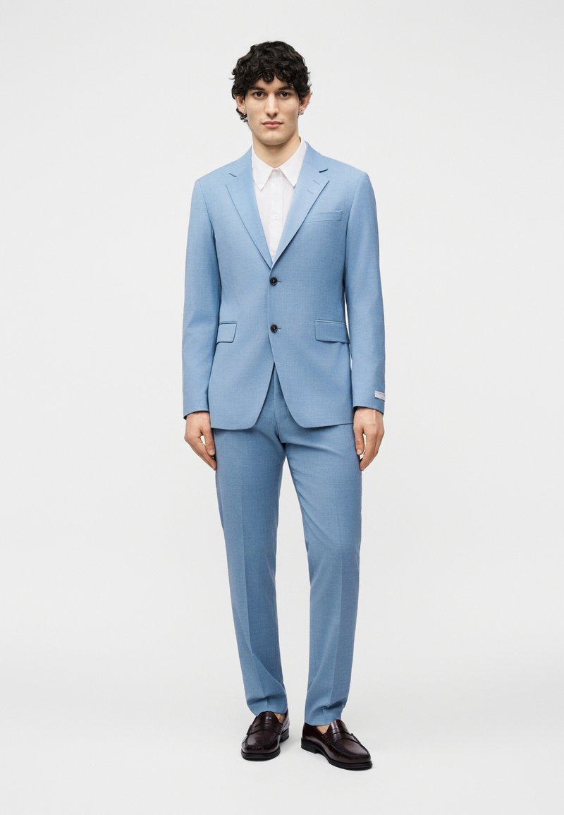 Άνδρας που στέκεται φορώντας ανοιχτό μπλε κοστούμι με μαύρα παπούτσια και λευκό πουκάμισο, κοιτάζοντας μπροστά σε απλό φόντο.