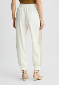 Pantalons de jogging blancs avec un panneau en maille dorée texturée sur les côtés. Présentent une taille froncée et des poignets ajustés aux chevilles.